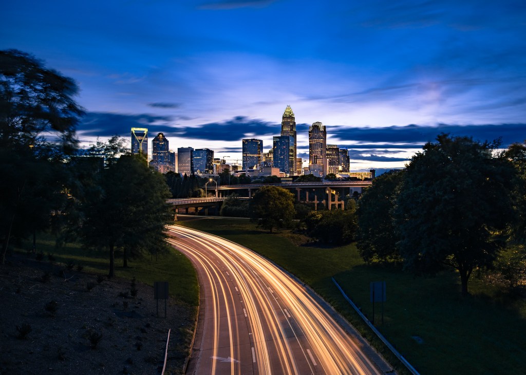 A long-exposure photo shows streaks of light from cars along a dark highway headed into the Charlotte skyline as the sun sets.