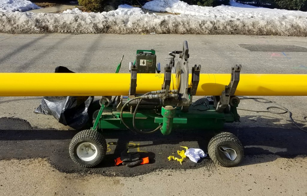 A green cart-like machine holds a yellow natural gas pipeline on the street.