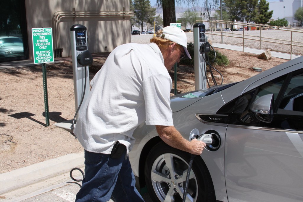 A man in a white shirt and baseball cap plugs in an electric vehicle in Las Vegas. The ground around him is a dustry red.